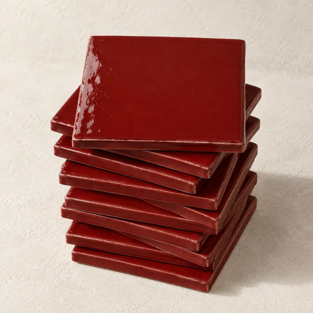 A stack of glossy square ceramic coasters in a deep scarlet red colour, arranged on a textured off-white surface.