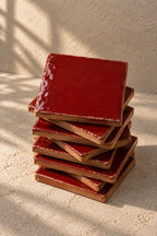 A stack of glossy square ceramic coasters in a deep scarlet red colour, arranged on a textured off-white surface.