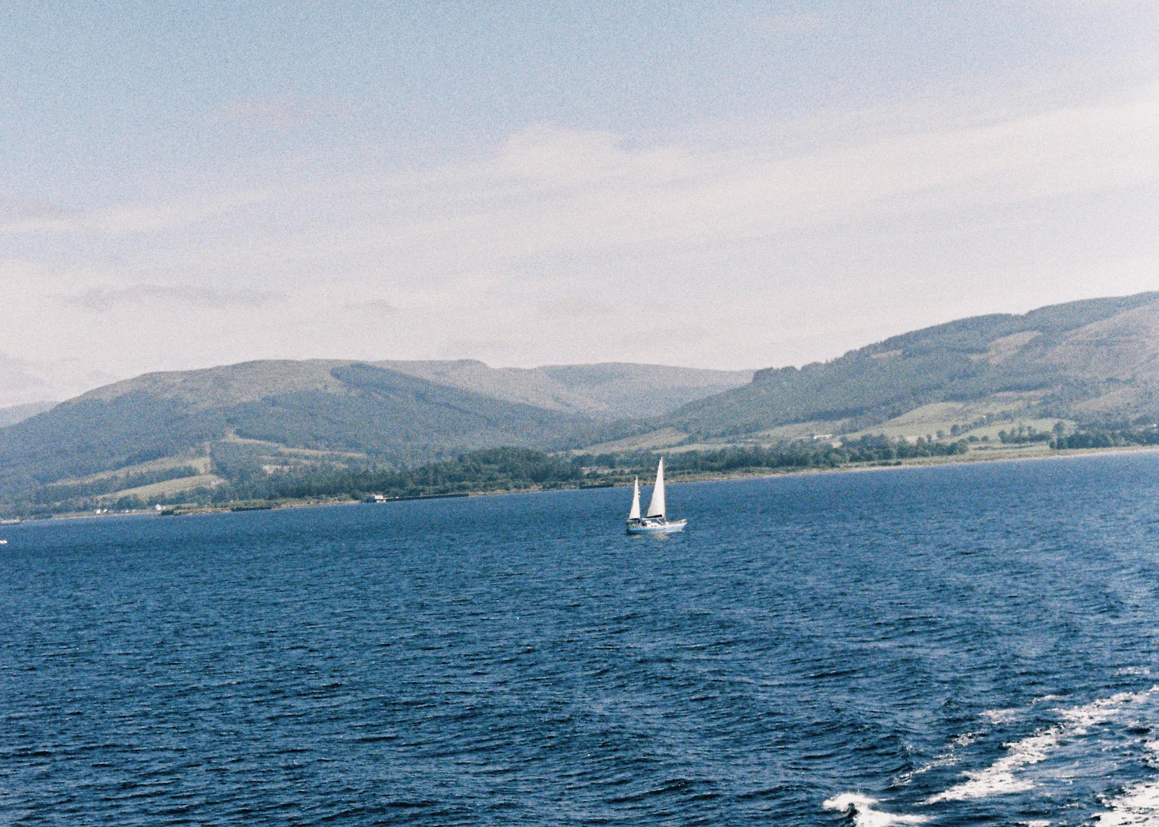 Film photograph of a lone sailboat on a deep blue loch with rolling green hills in the background — shot on 35mm film with a soft, hazy light.