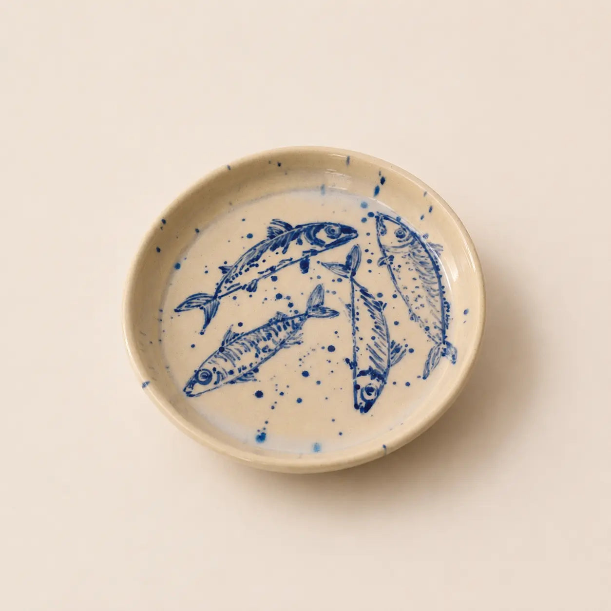A round ceramic small plate with a hand-painted blue fish design on a warm cream background, photographed from directly above on a matching cream surface.