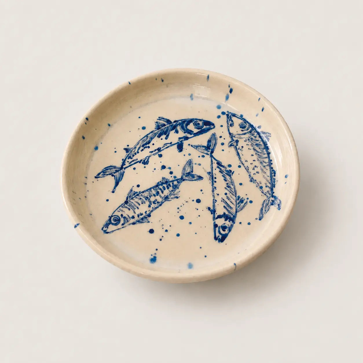 A round ceramic small plate with a hand-painted blue fish design on a warm cream background, photographed from directly above on a matching cream surface.
