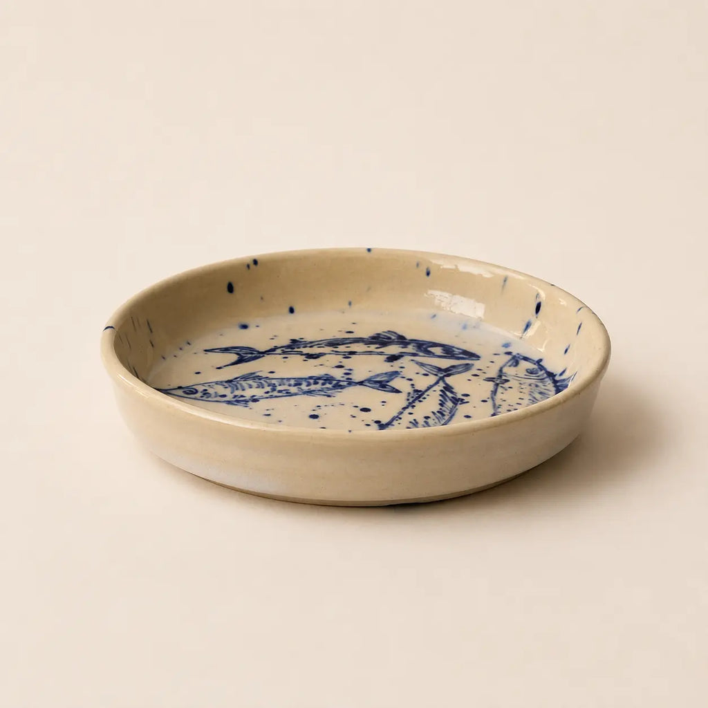 A shallow round ceramic small plate with a speckled beige exterior and a blue fish illustration printed inside, set against a solid cream background.