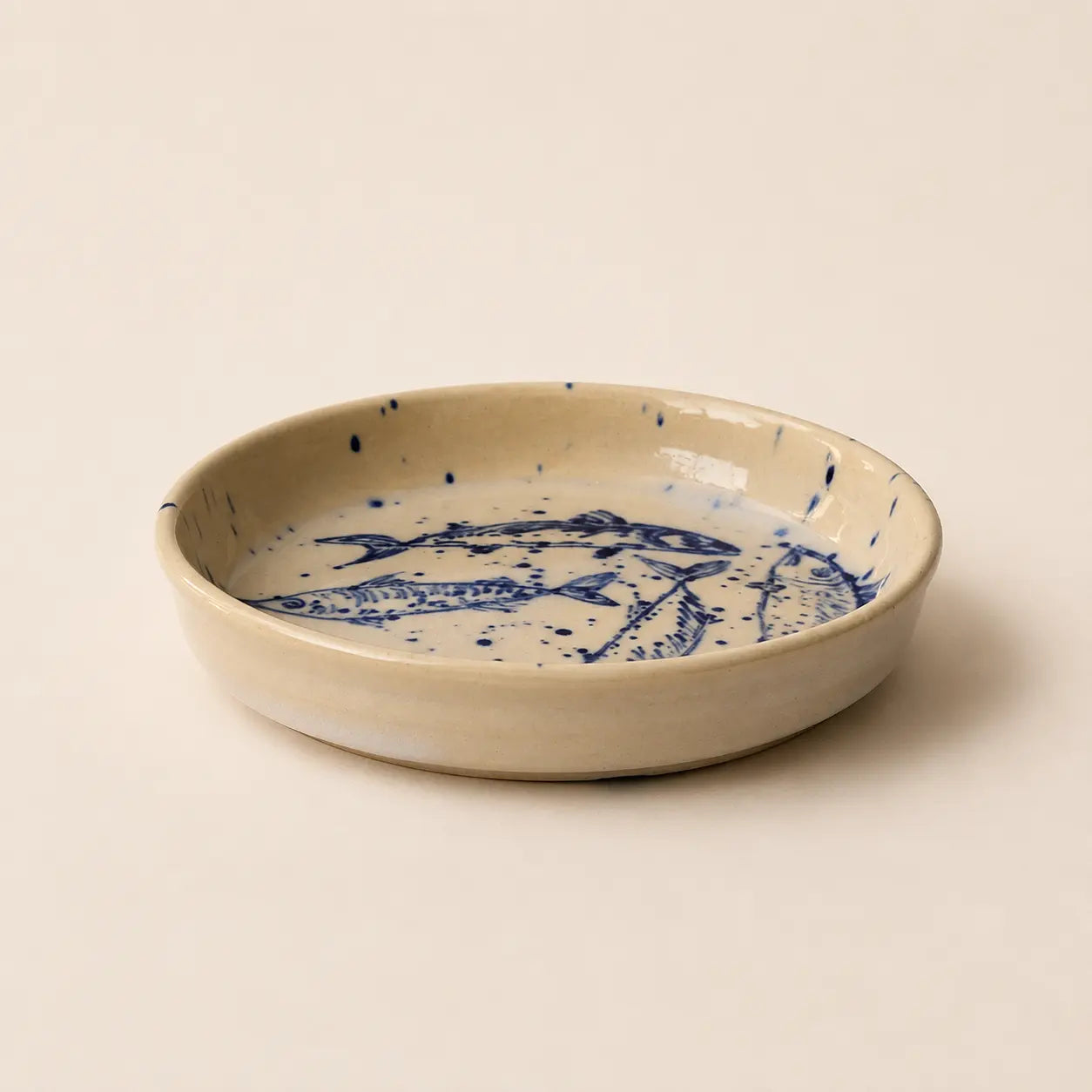 A shallow round ceramic small plate with a speckled beige exterior and a blue fish illustration printed inside, set against a solid cream background.