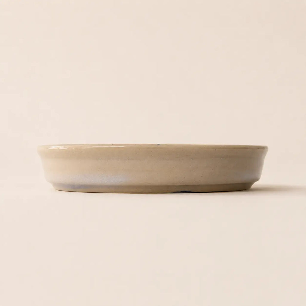 A shallow, wide ceramic small plate with a matte, light beige finish and a subtle cool-toned glaze along the bottom edge, set against a solid, warm off-white background.