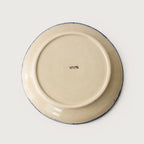 The underside of a round ceramic plate or dish, featuring a cream-colored glaze with a thin dark blue rim and a small black manufacturer's mark in the center, set against a plain light gray background.