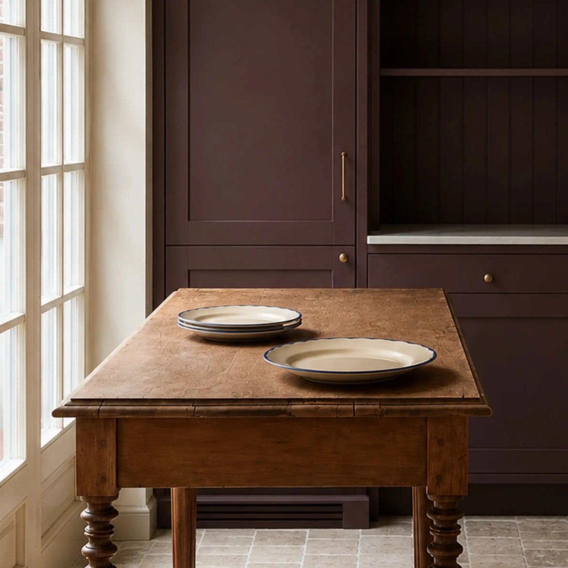 A rustic wooden dining table with two beige plates with dark blue rims, set in a kitchen with dark plum cabinets and a window on the left.