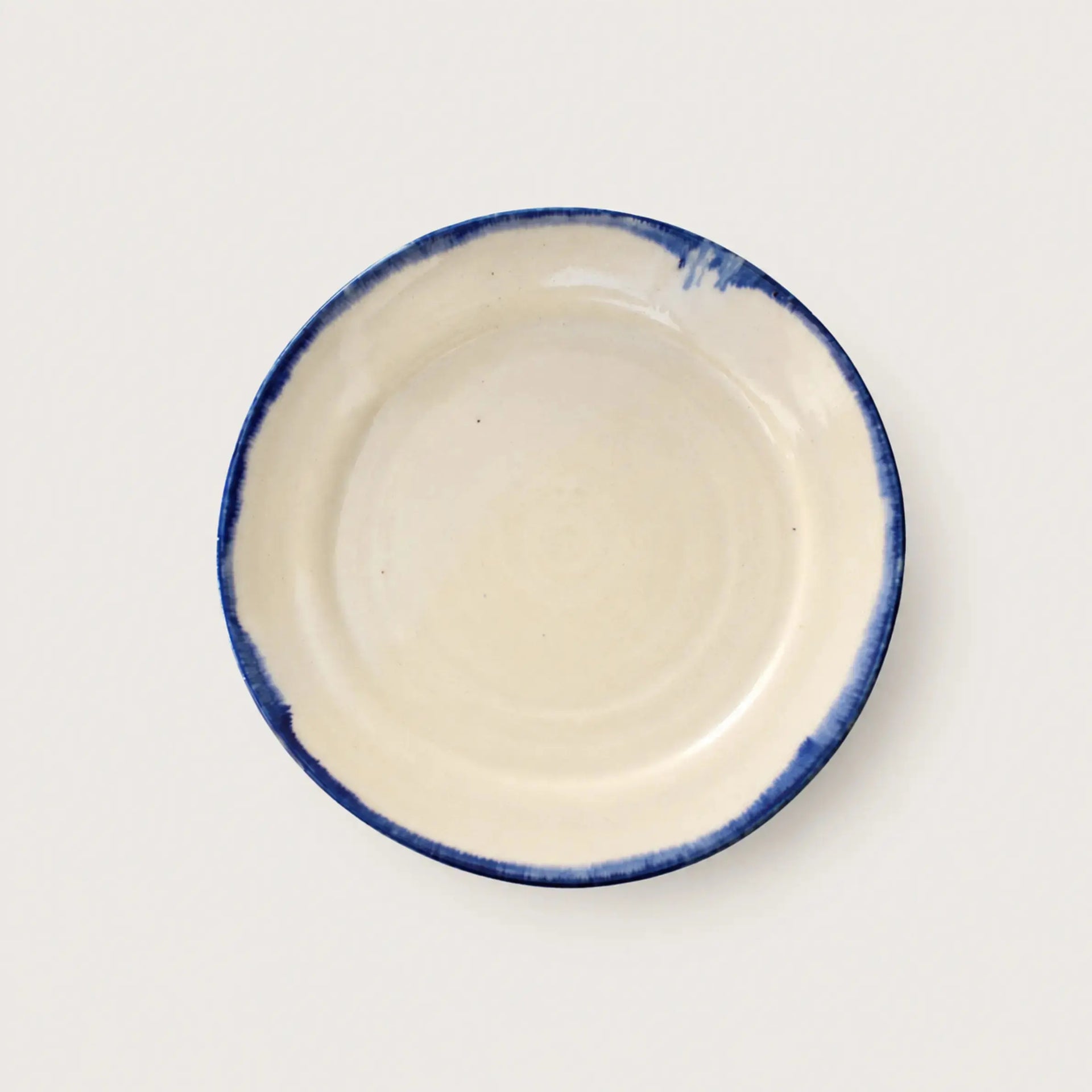 A round ceramic plate with a glossy off-white interior and a rustic, hand-painted dark blue rim, shown from a top-down perspective on a light gray background.