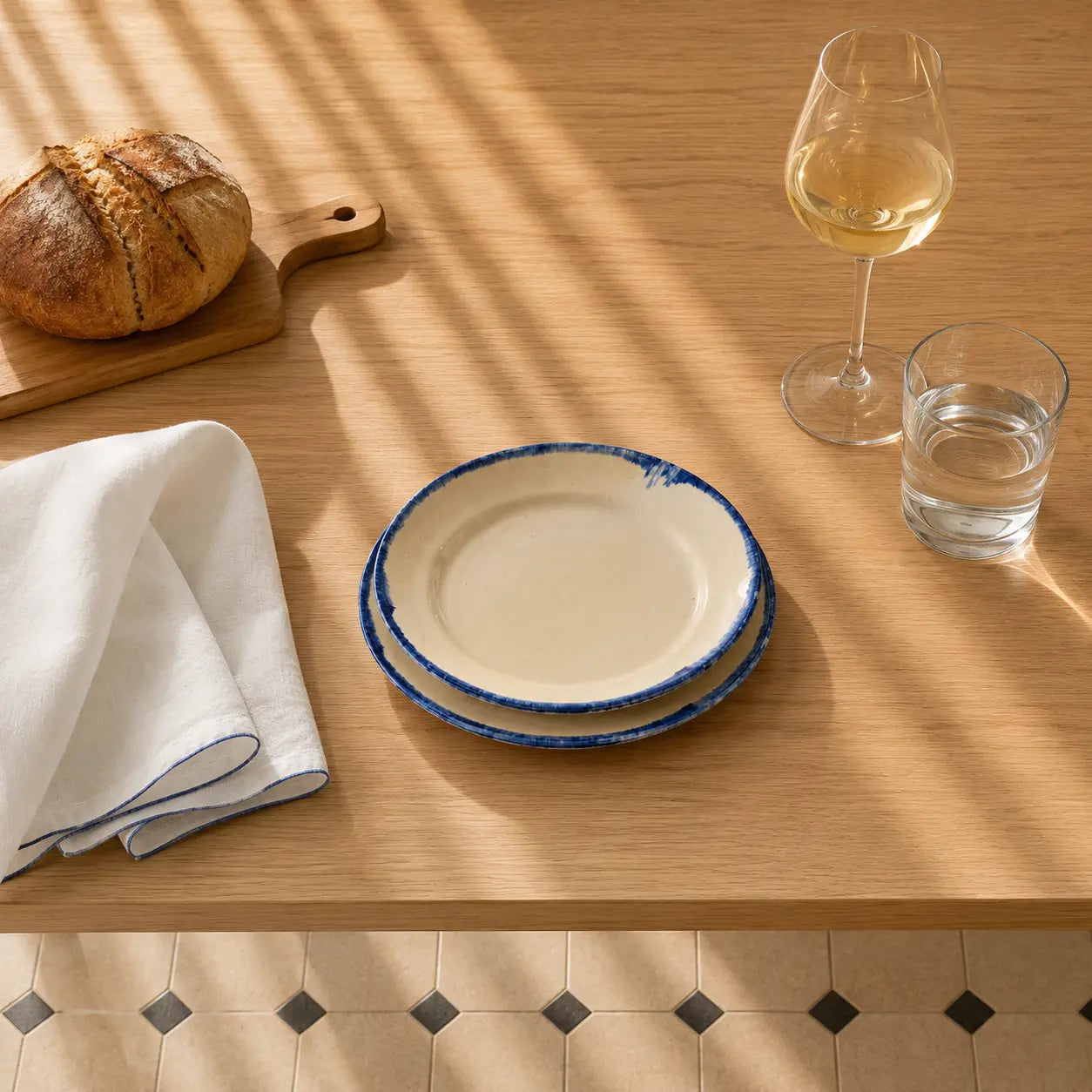 Two stacked cream-colored ceramic plates with a rustic blue rim, styled on a light wood table next to a folded white napkin, a loaf of bread on a wooden board, a glass of white wine, and a glass of water under soft window shadows.