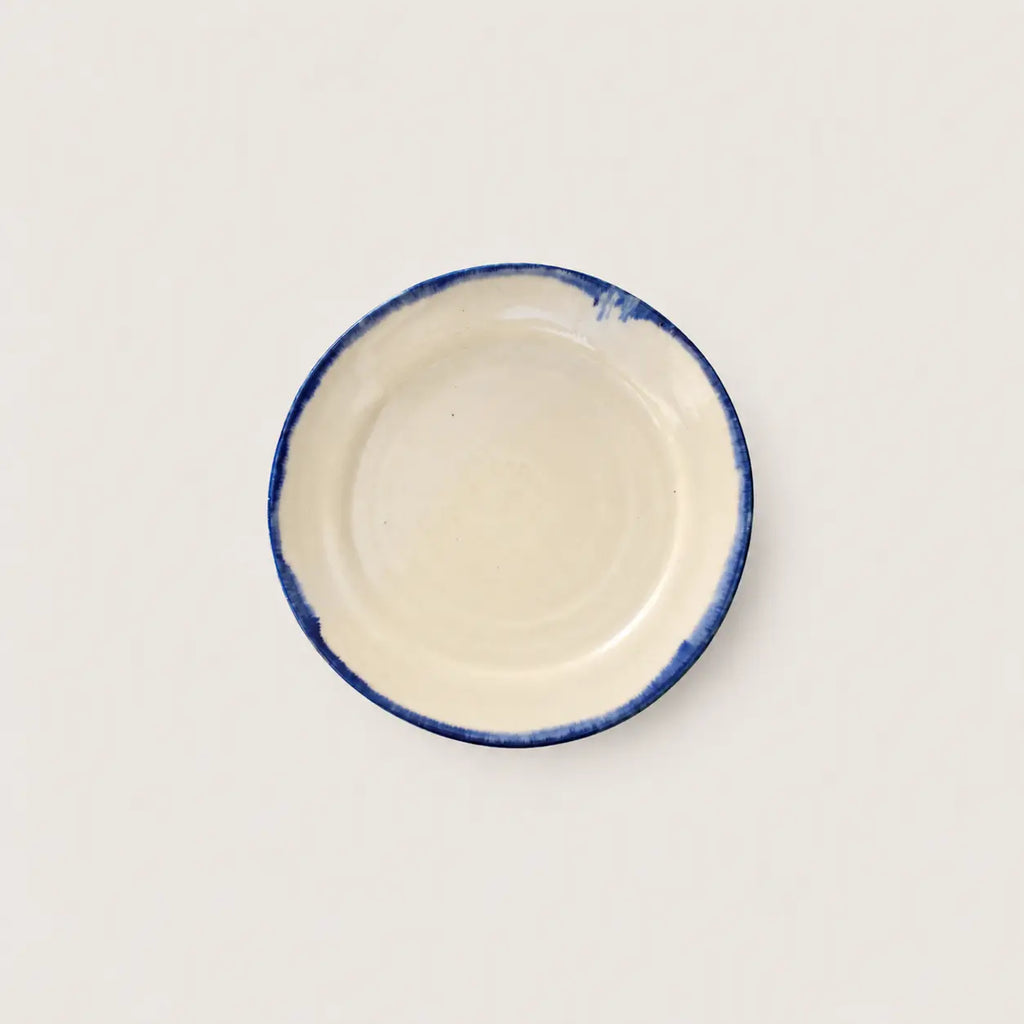 A small round ceramic side plate with a cream-colored interior and a hand-painted blue rim, shown from a top-down view on a light off-white background.