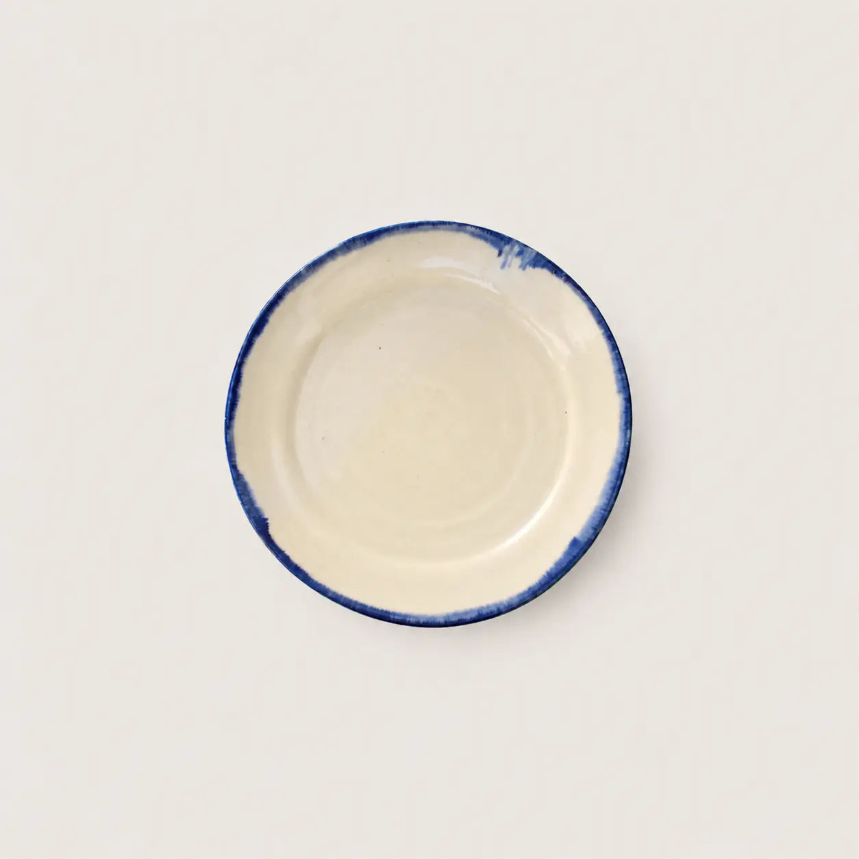 A small round ceramic side plate with a cream-colored interior and a hand-painted blue rim, shown from a top-down view on a light off-white background.