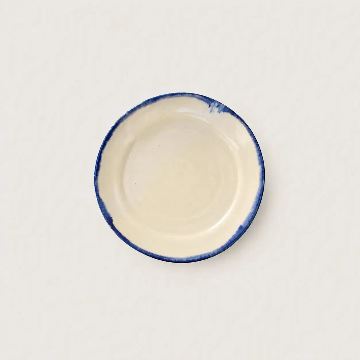 A small round ceramic side plate with a cream-colored interior and a hand-painted blue rim, shown from a top-down view on a light off-white background.