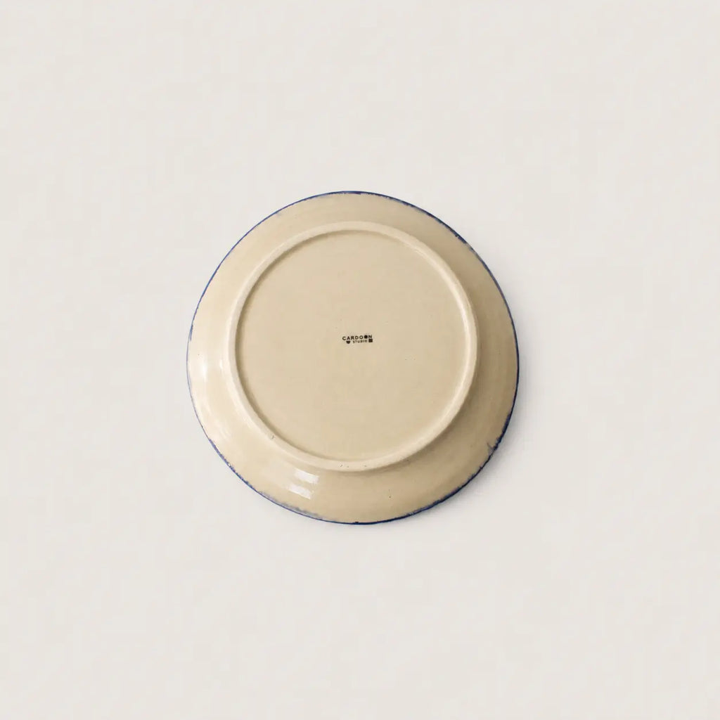 The underside of a round ceramic side plate or dish, showing a cream-coloured glaze with a thin dark blue rim and a small printed brand mark in the centre, set against a plain off-white background.