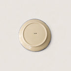 The underside of a round ceramic side plate or dish, showing a cream-coloured glaze with a thin dark blue rim and a small printed brand mark in the centre, set against a plain off-white background.