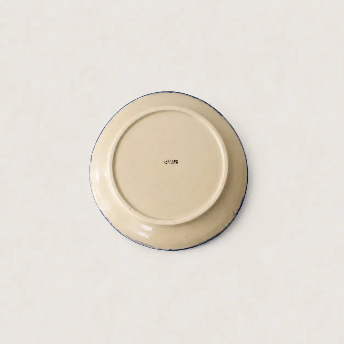 The underside of a round ceramic side plate or dish, showing a cream-coloured glaze with a thin dark blue rim and a small printed brand mark in the centre, set against a plain off-white background.