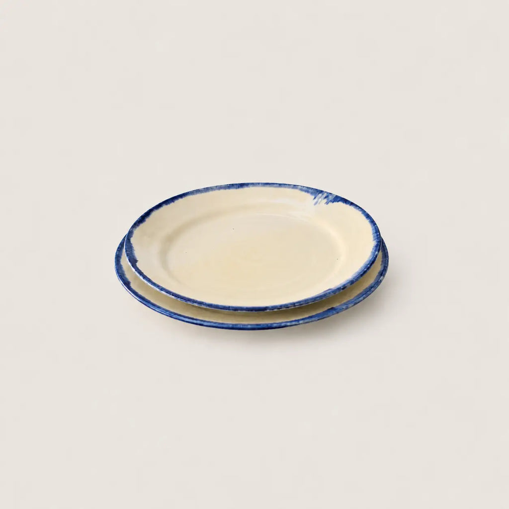 Two small round side plates stacked on top of each other, featuring a warm cream-colored glaze and a rustic dark blue hand-painted rim, set against a solid light beige background.