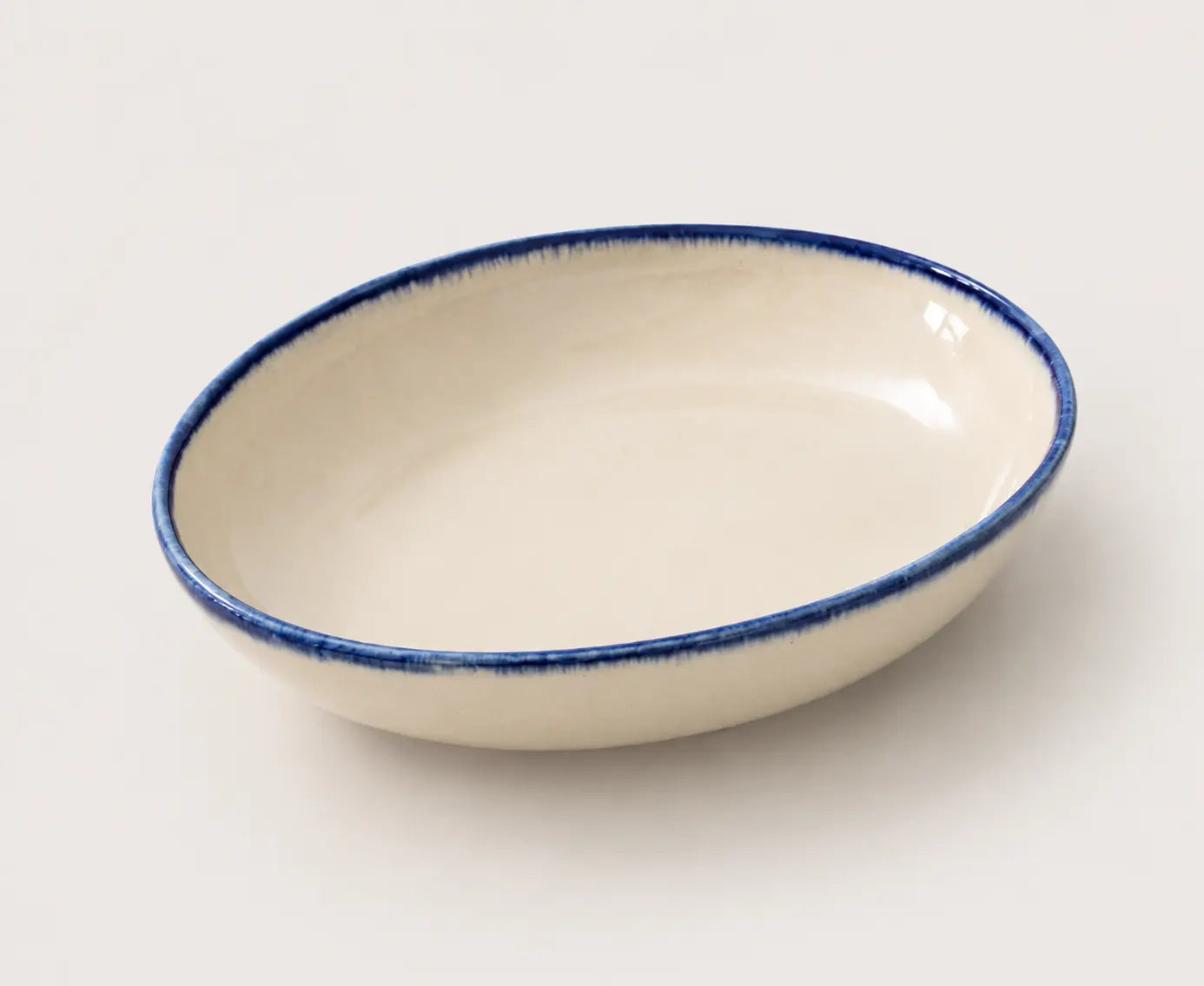 An oval ceramic soup bowl with a glossy off-white interior and a textured, hand-painted dark blue rim, shown on a neutral light gray background.
