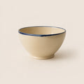 A small ceramic soup bowl with a warm cream-colored interior and exterior, featuring a contrasting dark blue hand-painted rim, set against a solid cream background.