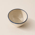 A small handmade ceramic soup bowl with a cream-colored interior and exterior, featuring a contrasting dark blue painted rim, set against a solid light beige background.