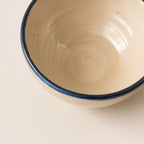 A close-up of a ceramic soup bowl's interior, featuring a warm cream-coloured glaze with a contrasting dark blue rim, set against a matching cream background.