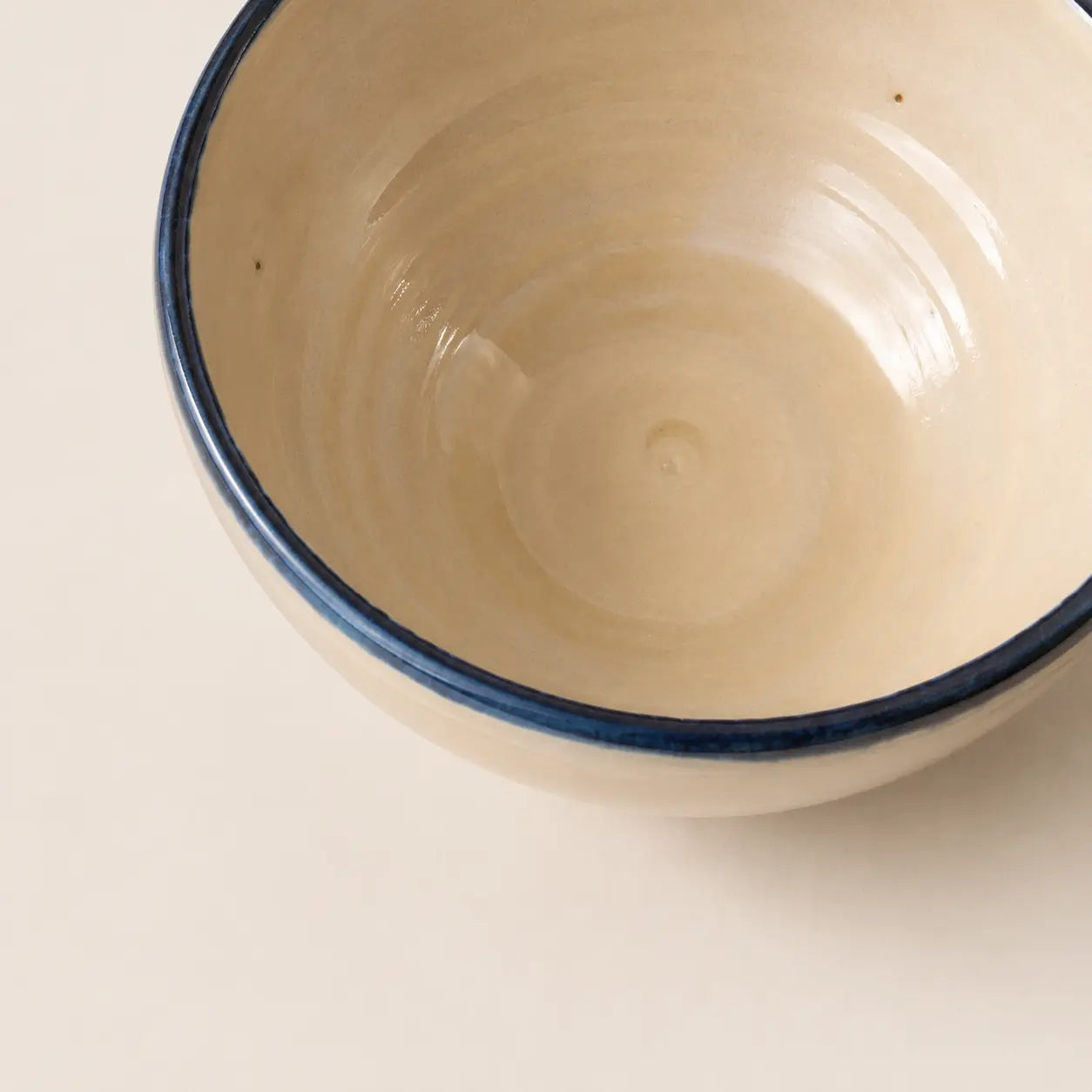 A close-up of a ceramic soup bowl's interior, featuring a warm cream-coloured glaze with a contrasting dark blue rim, set against a matching cream background.