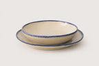 A ceramic soup bowl with a matching dinner plate, both in a warm cream-beige glaze with a rustic dark blue rim, set against a plain light gray background.