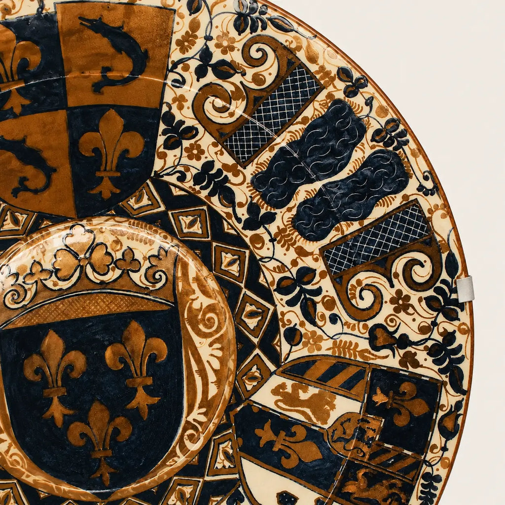 A close-up of an ornate round decorative plate or tray featuring a detailed heraldic pattern in navy blue, gold, and cream, with fleur-de-lis motifs and shield-like sections.