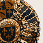 A close-up of an ornate round decorative plate or tray featuring a detailed heraldic pattern in navy blue, gold, and cream, with fleur-de-lis motifs and shield-like sections.