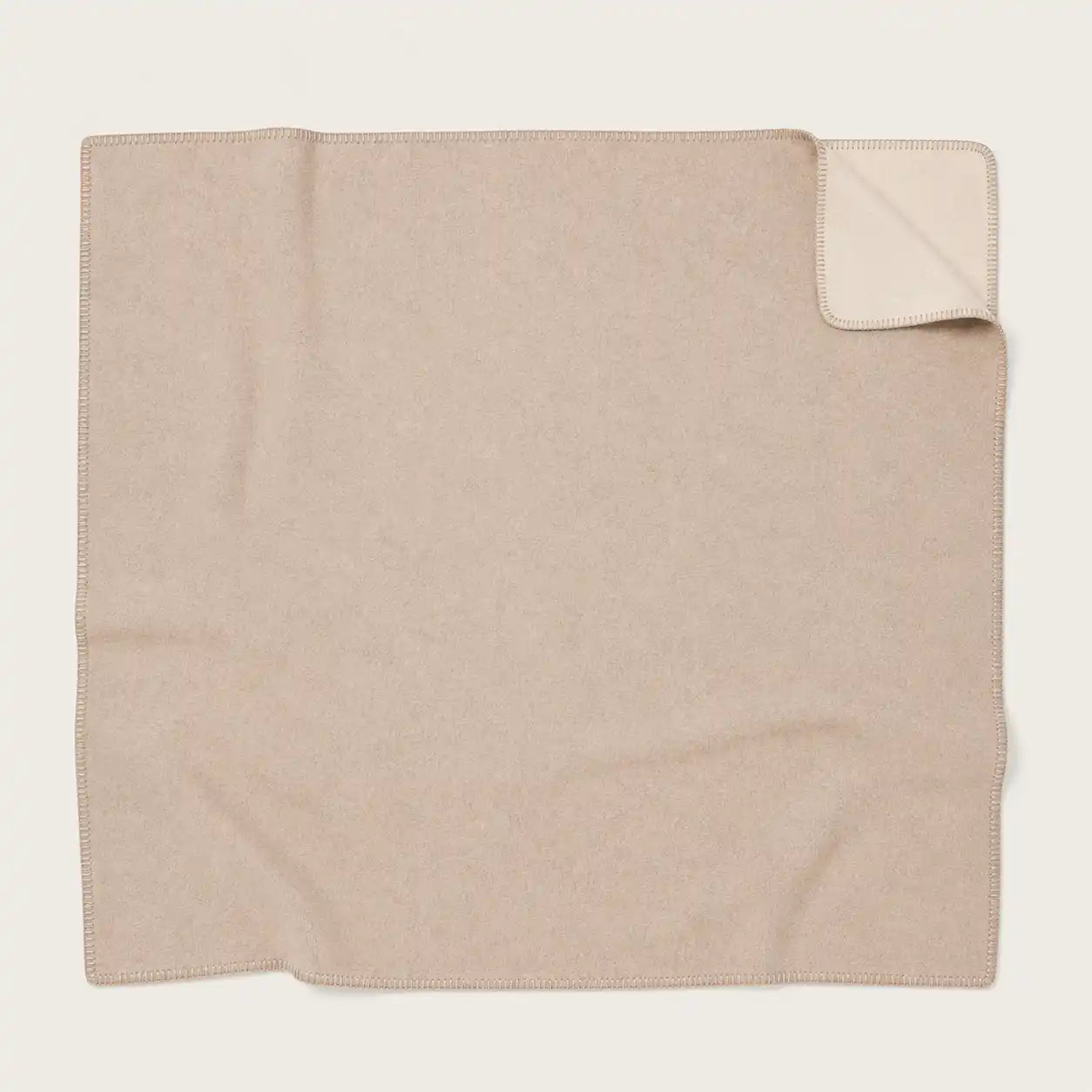 A folded beige throw blanket with a small corner turned up to show the reverse side, photographed on a plain light background.