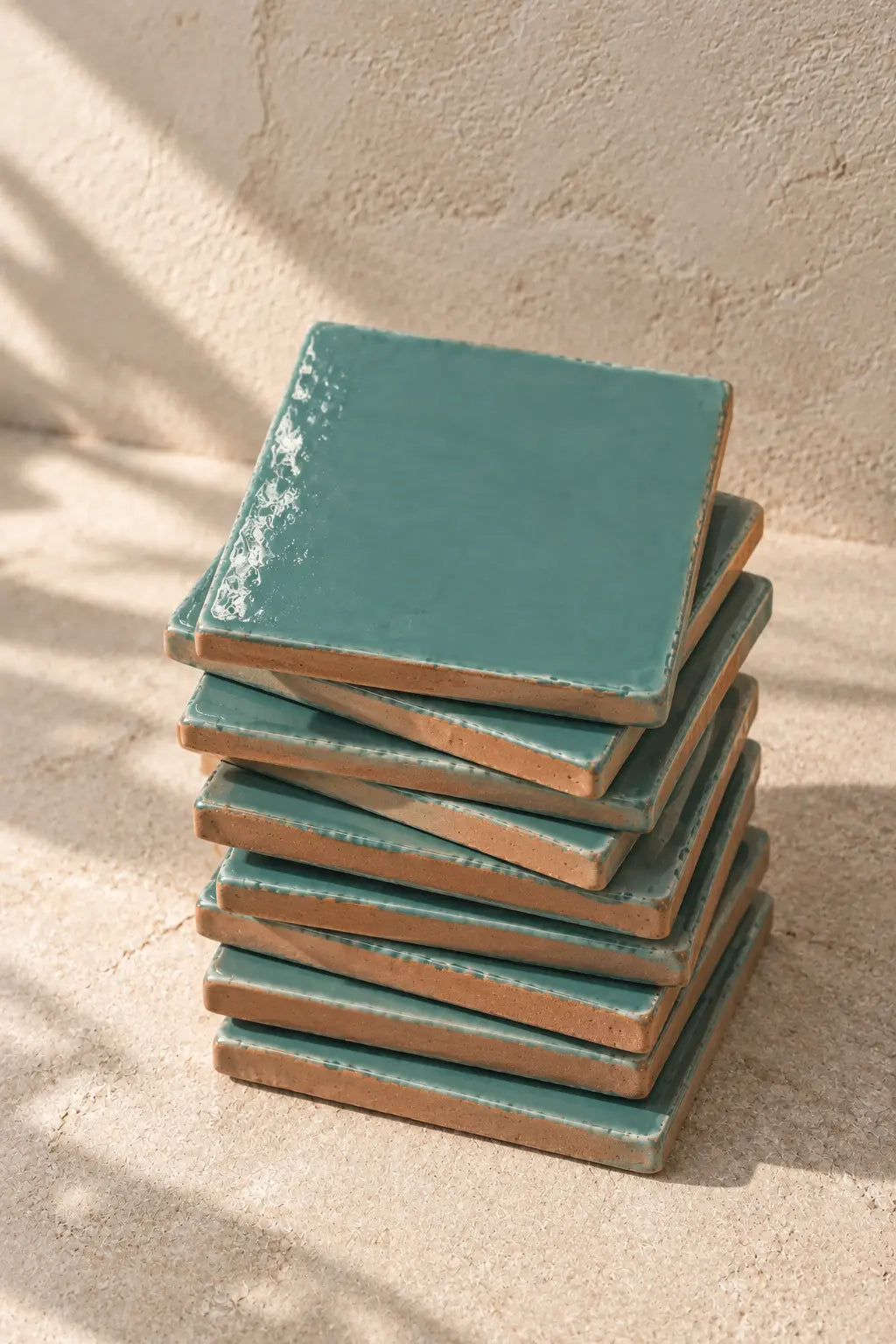 A stack of square ceramic coasters in a glossy sky blue glaze, set against a textured beige wall and surface.