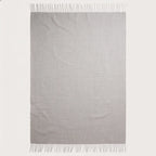 A light gray woven throw blanket with fringed edges, displayed flat against a solid off-white background.