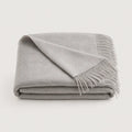 A folded light gray throw blanket with fringed edges, displayed on a clean off-white background.