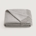 A folded light gray throw blanket with fringed edges, displayed on a clean off-white background.