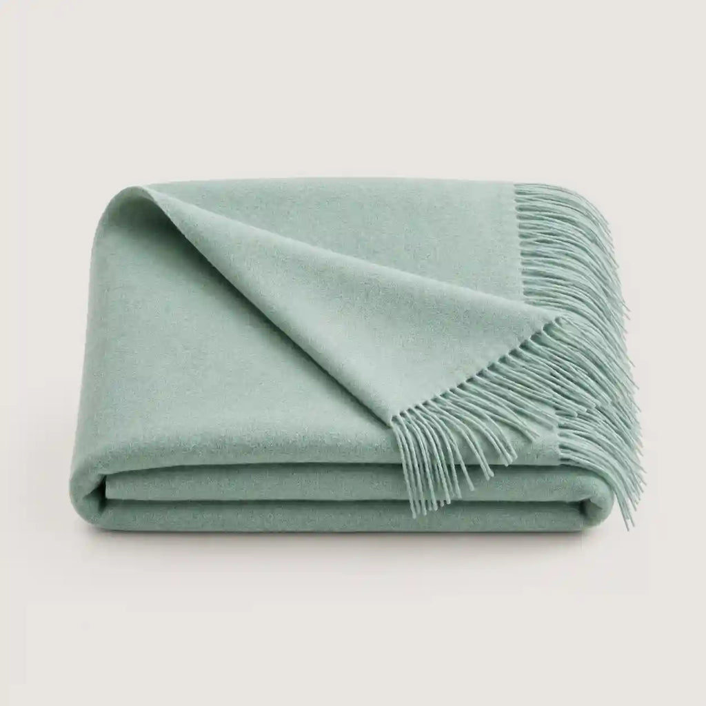 A folded sage green throw blanket with fringed edges, displayed on a clean, light off-white background.