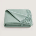 A folded sage green throw blanket with fringed edges, displayed on a clean, light off-white background.