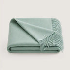 A folded sage green throw blanket with fringed edges, displayed on a clean, light off-white background.