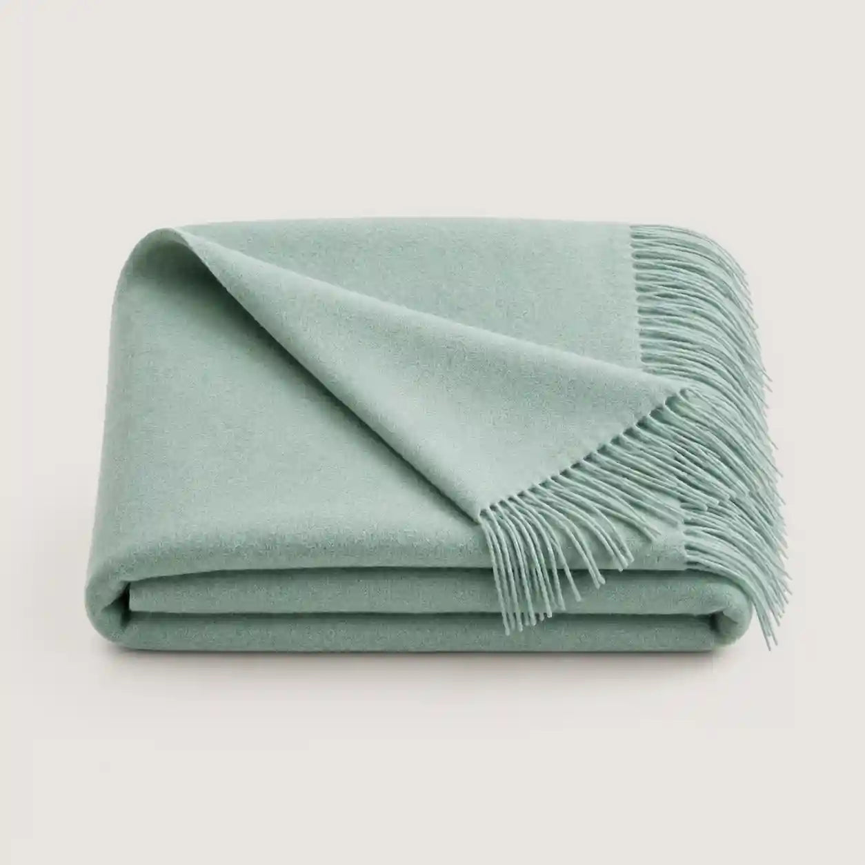 A folded sage green throw blanket with fringed edges, displayed on a clean, light off-white background.