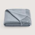 A folded light blue throw blanket with fringed edges, displayed on a clean, solid, light off-white background.