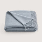 A folded light blue throw blanket with fringed edges, displayed on a clean, solid, light off-white background.
