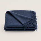 A folded navy blue throw blanket with fringed edges, displayed on a clean, light beige background.