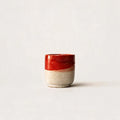 A small handmade ceramic espresso cup with a glossy red glazed top half and a textured, speckled beige clay bottom half, set against a plain off-white background.