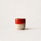 A small handmade ceramic espresso cup with a glossy red glazed top half and a textured, speckled beige clay bottom half, set against a plain off-white background.
