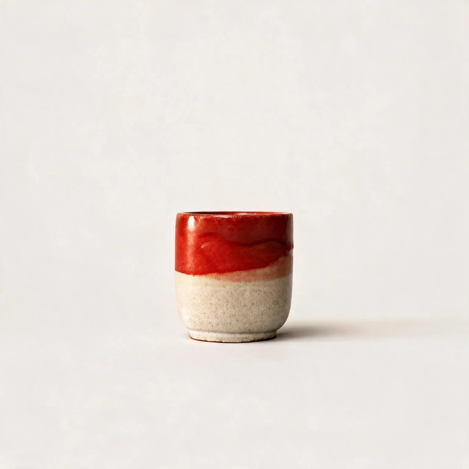 A small handmade ceramic espresso cup with a glossy red glazed top half and a textured, speckled beige clay bottom half, set against a plain off-white background.