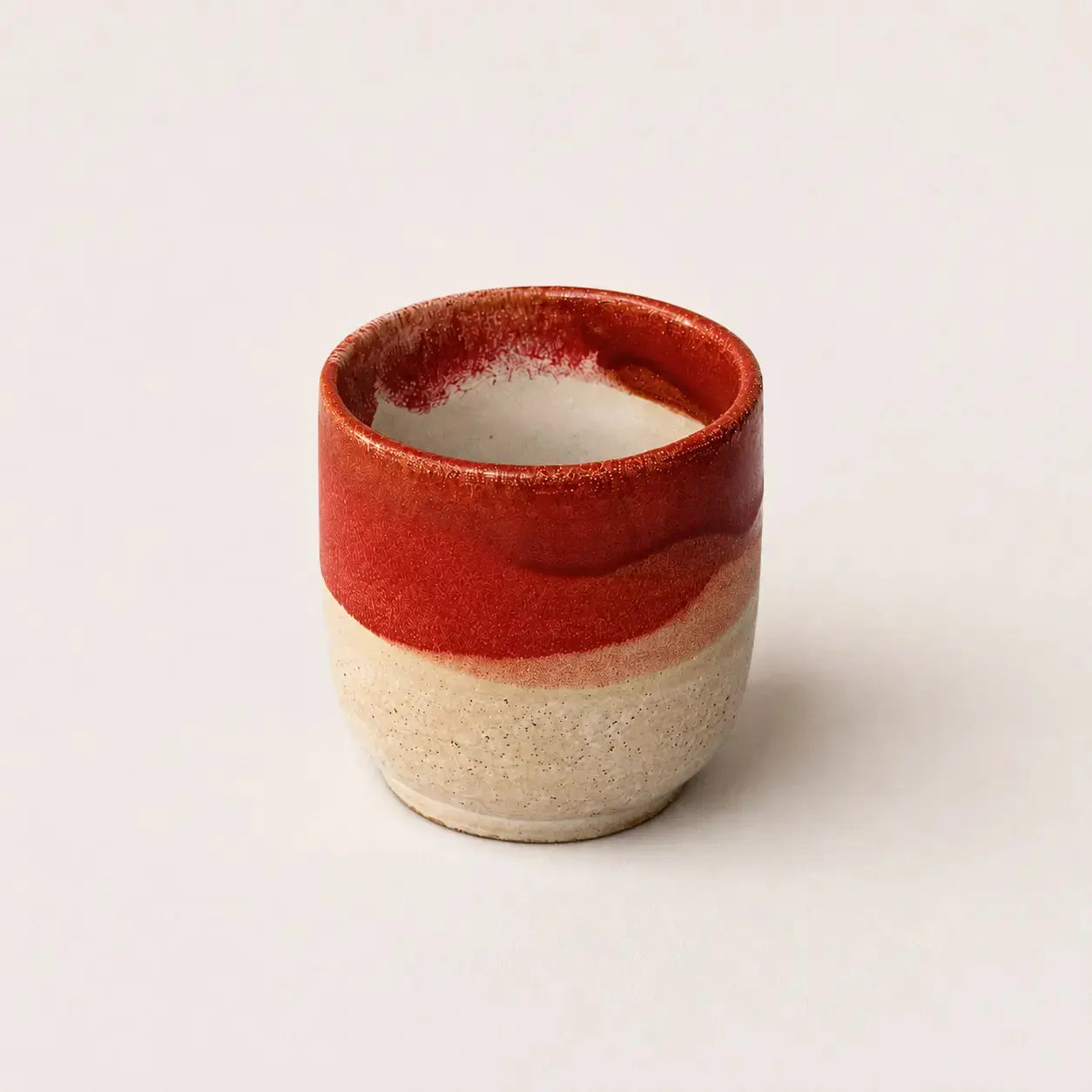 A small handmade ceramic espresso cup with a glossy red glazed upper half and a textured, speckled beige clay lower half, set against a plain light background.