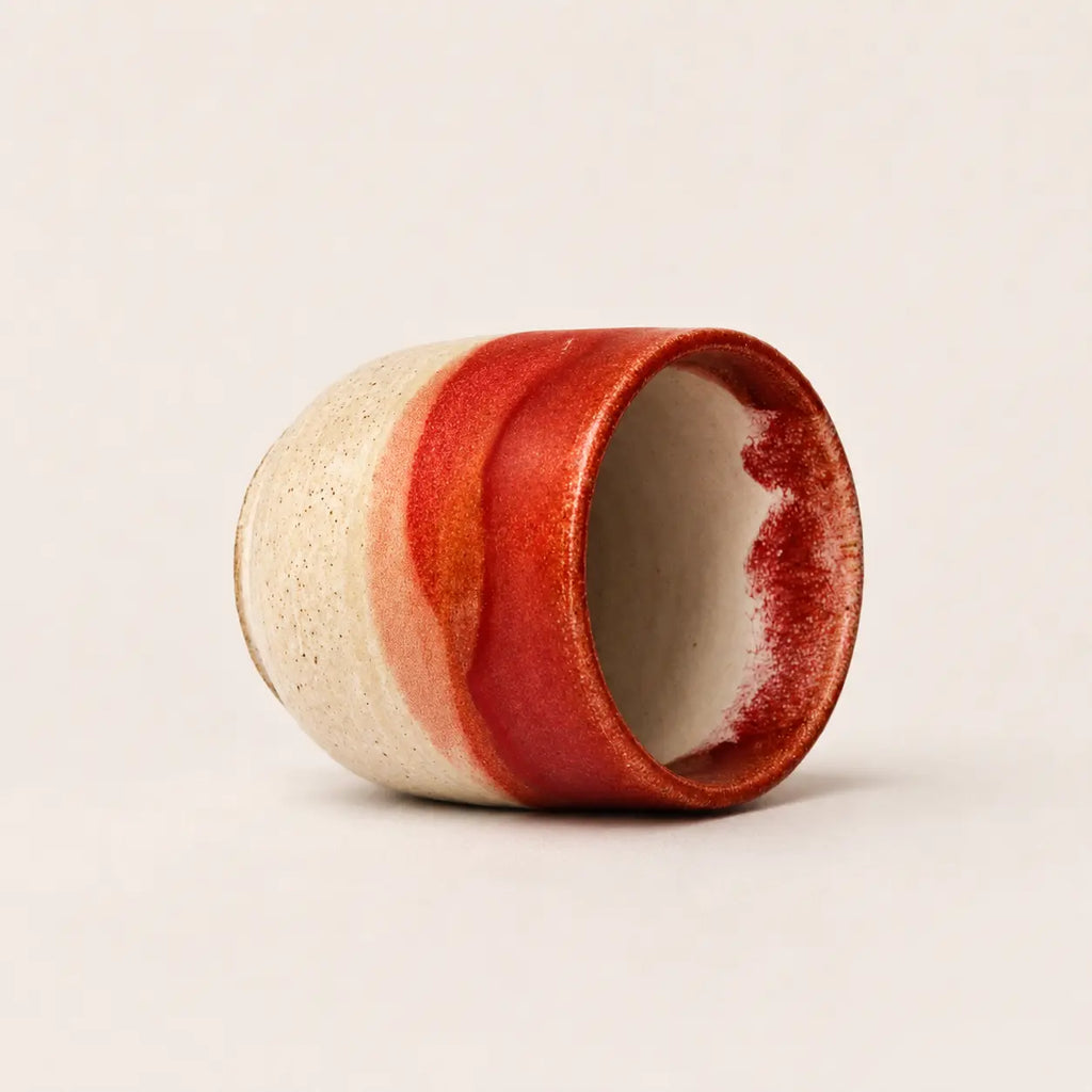 A handmade ceramic espresso cup with a textured cream exterior and a glossy red glazed finish, shown on a neutral light beige background.