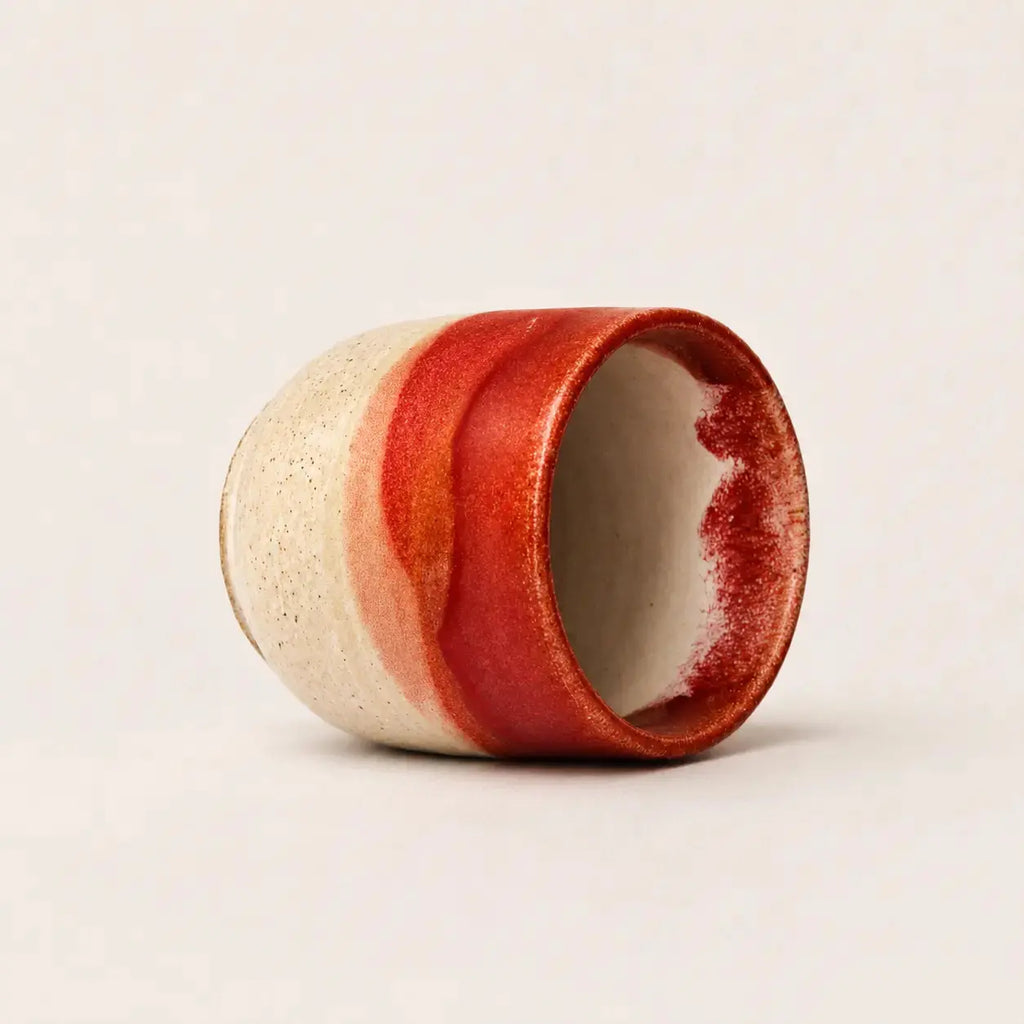 A handmade ceramic espresso cup with a textured cream exterior and a glossy red glazed finish, shown on a neutral light beige background.