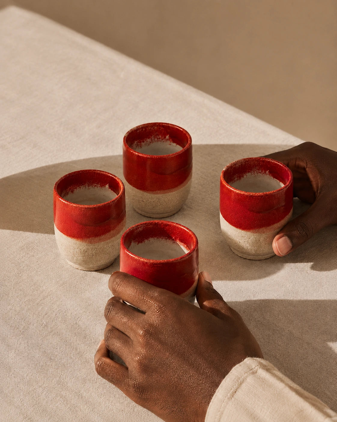 Four small ceramic cups with a glossy red and matte beige glaze are arranged on a light fabric surface, with two hands holding two of the cups.