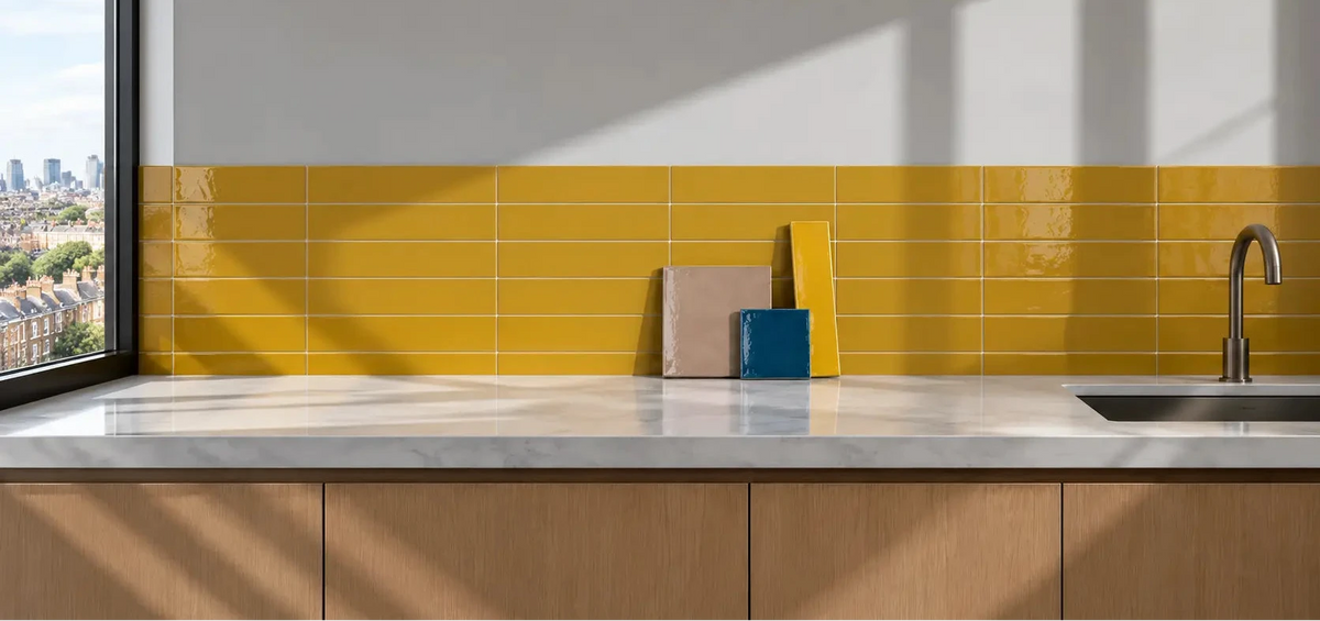 A modern kitchen backsplash featuring glossy yellow rectangular tiles across a white wall, with a white countertop, light wood cabinets, a chrome faucet, and a large window showing a city view.