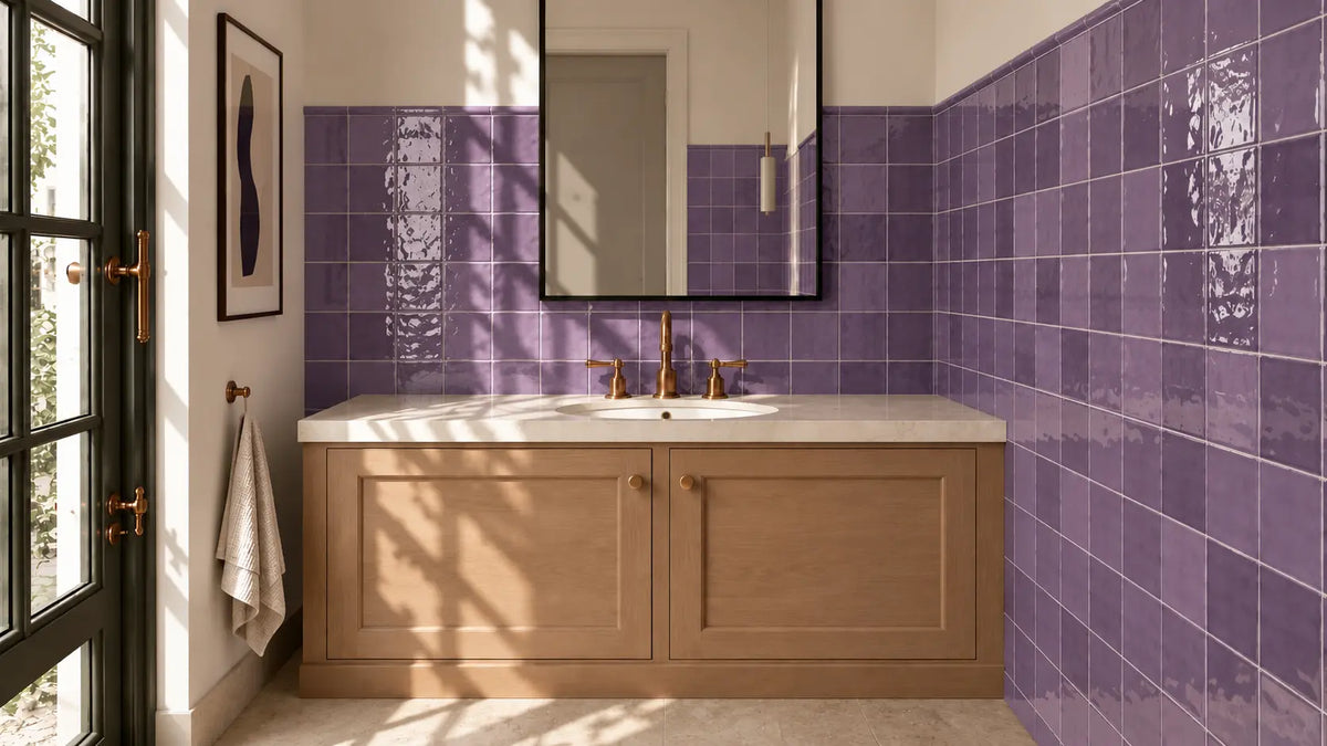 A modern bathroom vanity setup with glossy purple square wall tiles, a light wood cabinet, a white countertop, and a large mirror. Sunlight streams in from a window on the left, casting shadows across the vanity.