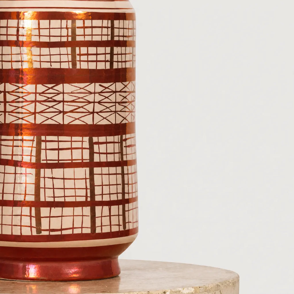 A close-up of a ceramic vase with a glossy copper-brown and cream geometric grid pattern, sitting on a round stone pedestal against a light gray background.