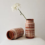 Two patterned ceramic vases in a terracotta and cream color scheme, one standing upright holding a white daisy and the other lying on its side, set against a neutral light gray background.